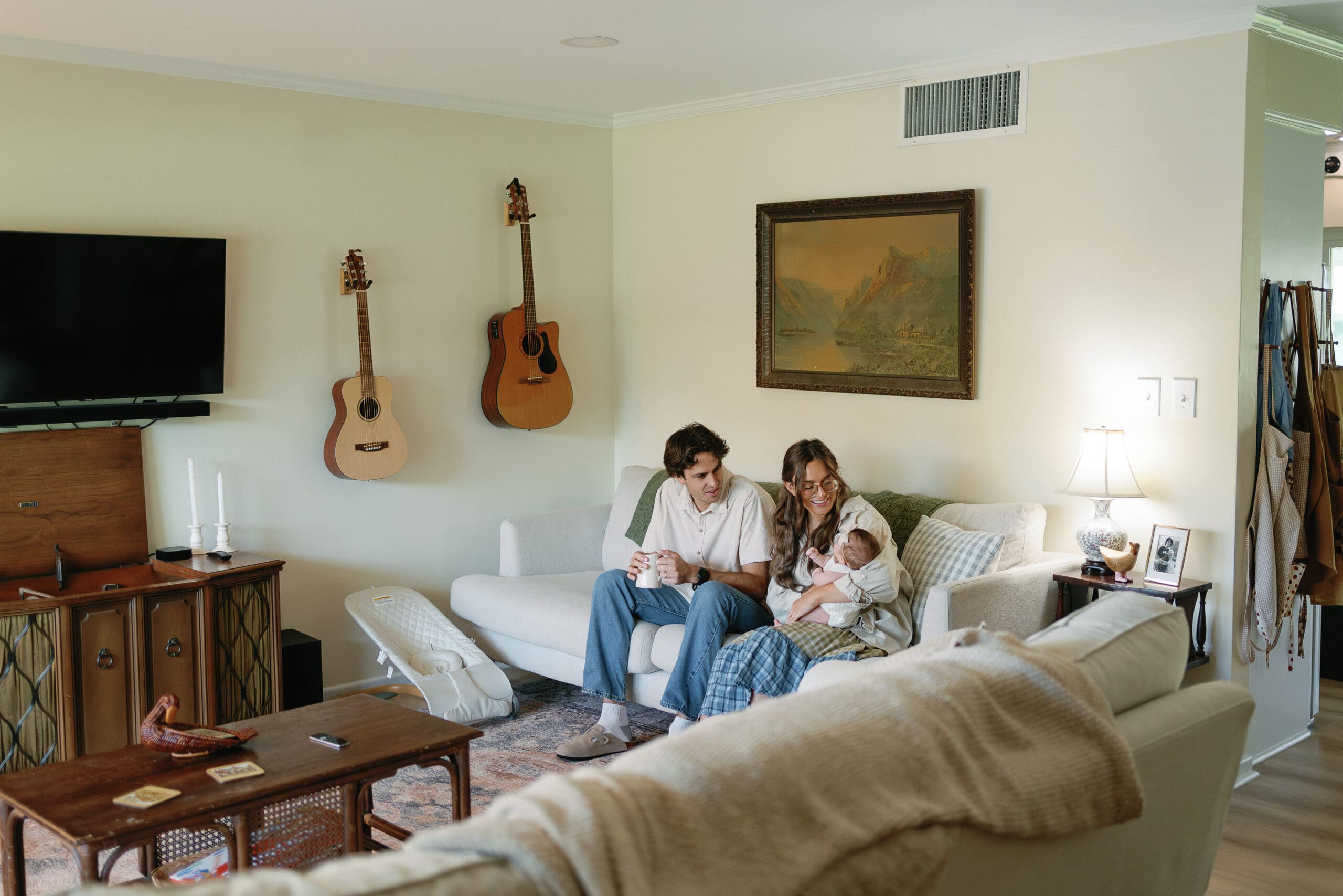 Parents sitting on couch in living room drinking coffee and looking at their baby